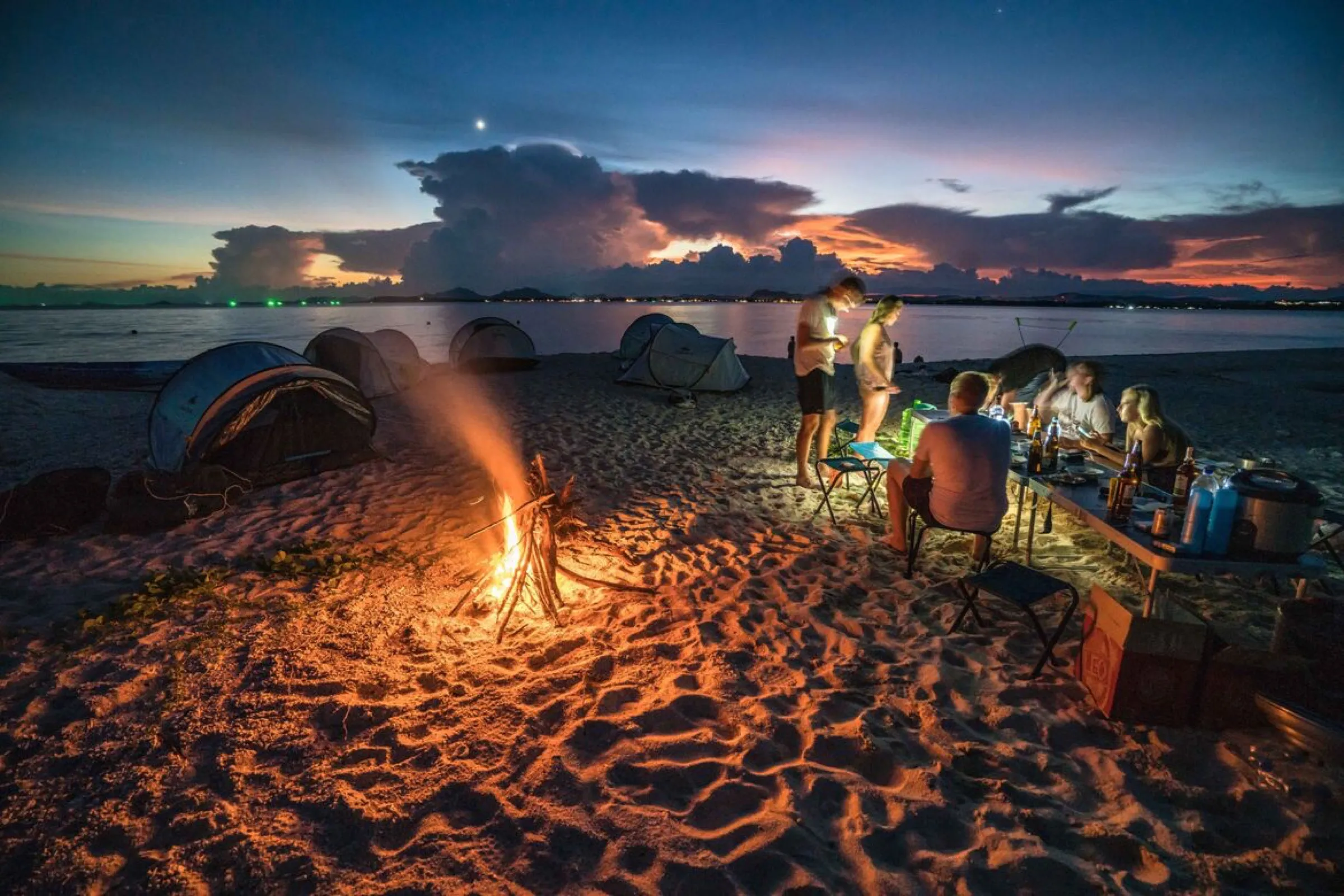 The group gathered around a campfire on the beach at dusk, tents behind, dramatic sky above.