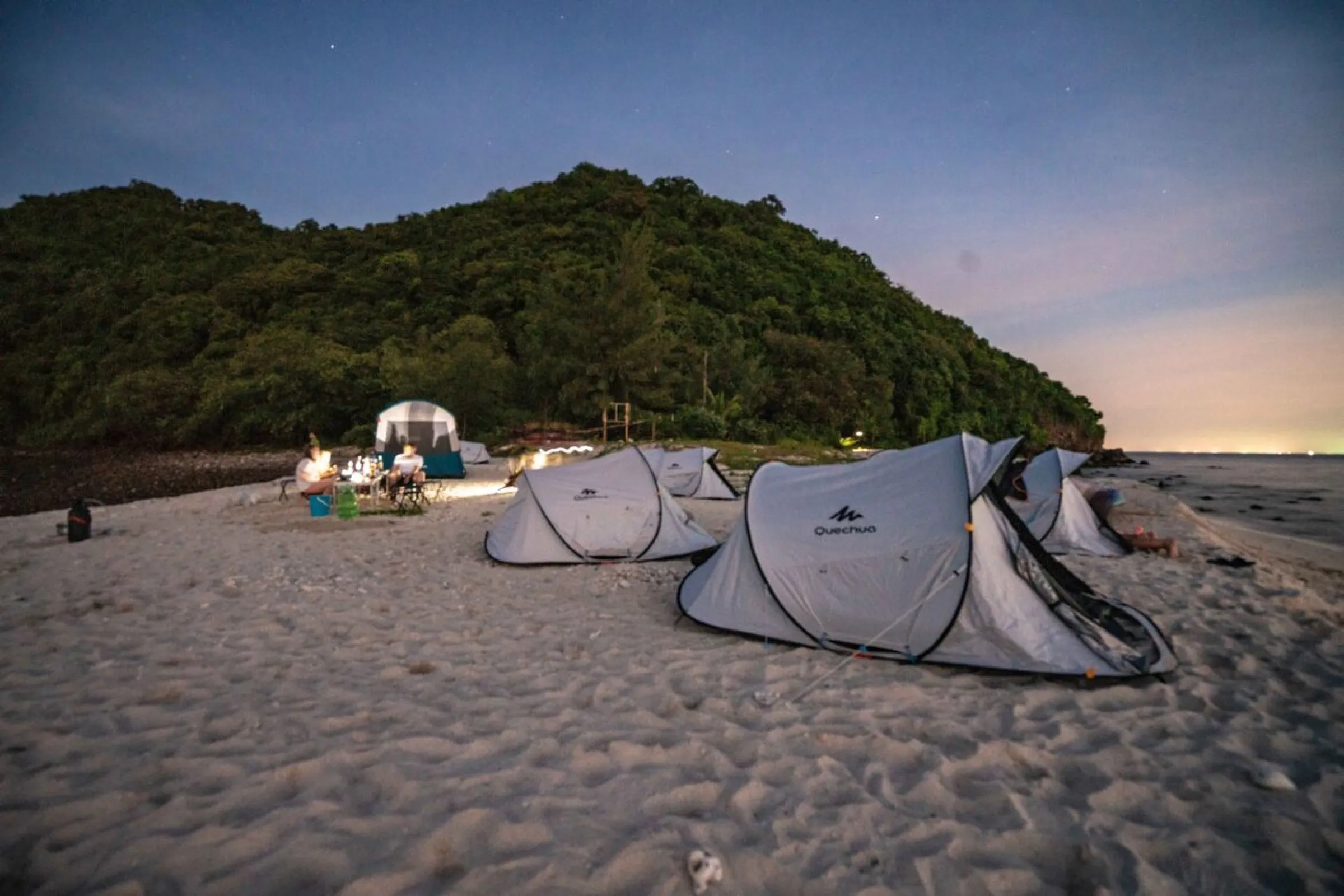 Tents glowing on the beach beneath a jungle hillside at twilight.