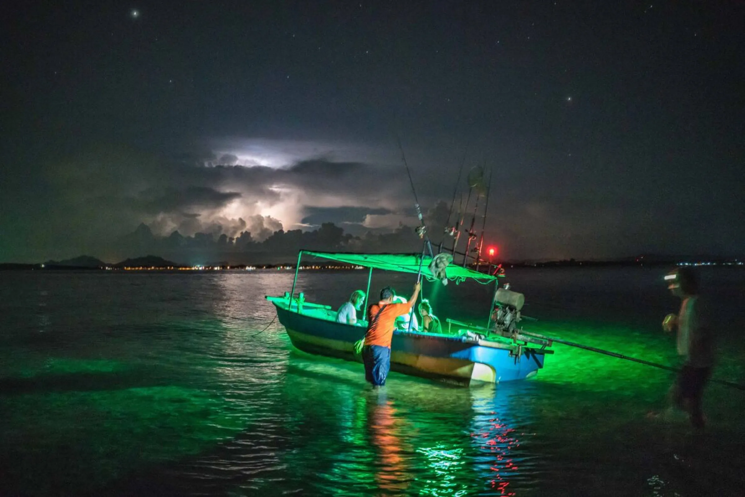 A traditional boat lit green over the water at night as a storm flickers on the horizon.
