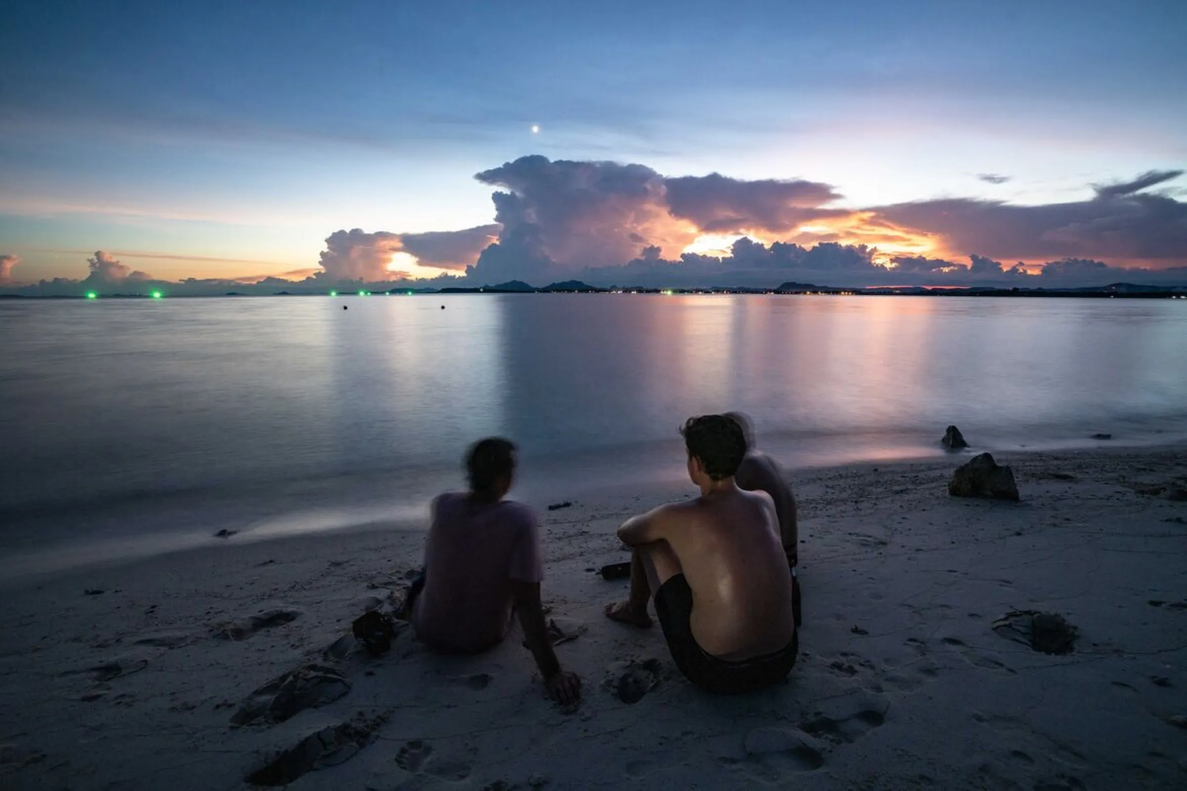 Two friends sitting on the sand watching the sun set over the calm bay.